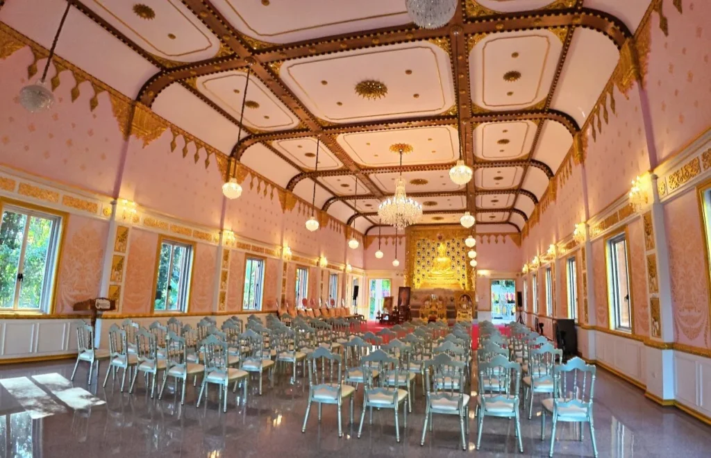 บรรยากาศภายในพระอุโบสถวัดจุฬาภรณ์วนาราม โทนสีชมพูทอง ประดับโคมไฟระย้า และจัดเก้าอี้รองรับผู้มาทำบุญ