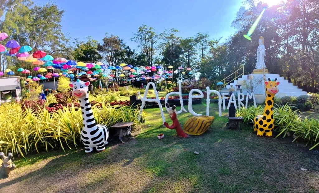 ป้าย Eden Farm นครนายก พร้อมรูปปั้นยีราฟและม้าลาย ท่ามกลางสวนสีเขียวและร่มหลากสีแขวนเต็มท้องฟ้า