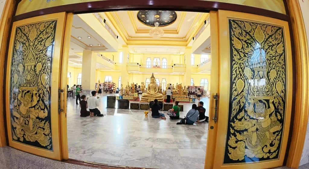 ประตูวิหารลวดลายไทยสีทอง เปิดสู่ภายในที่มีพระพุทธรูปและผู้คนสักการะ