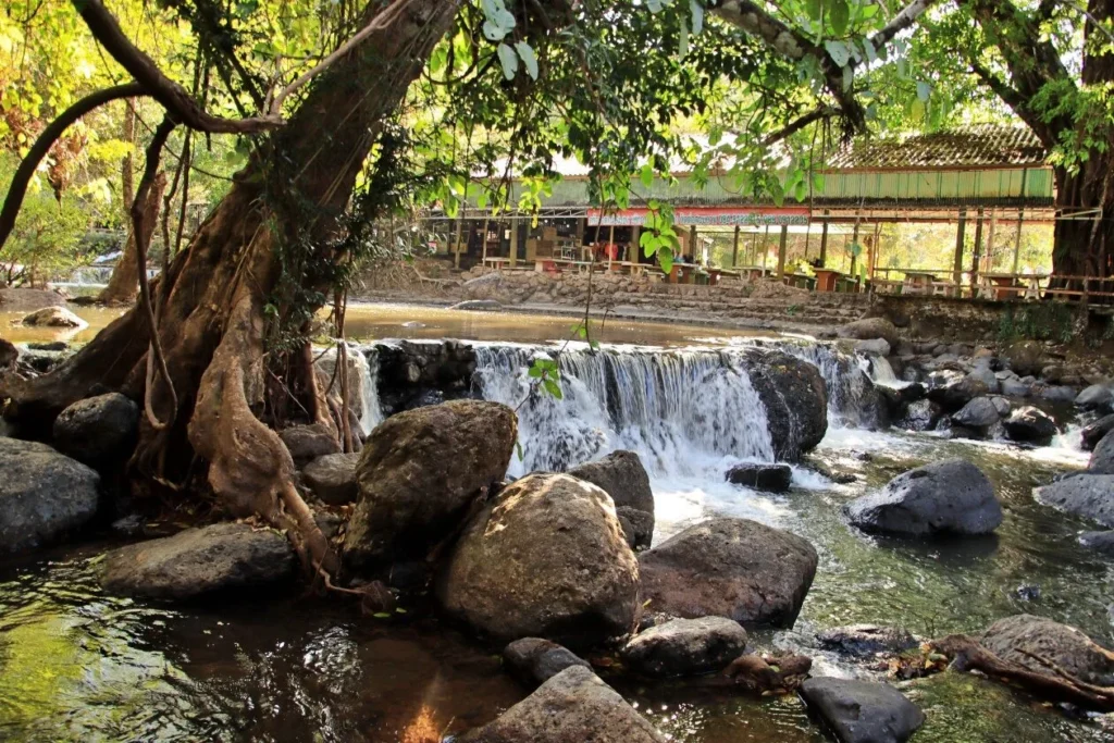 น้ำตกนางรอง จังหวัดนครนายก สายน้ำไหลผ่านโขดหินท่ามกลางธรรมชาติและร้านอาหารริมน้ำ
